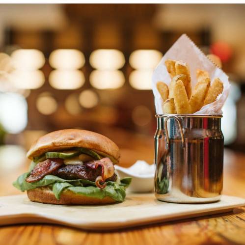 A delicious burger with bacon, lettuce, and pickles, served with a side of crispy fries in a metal cup.