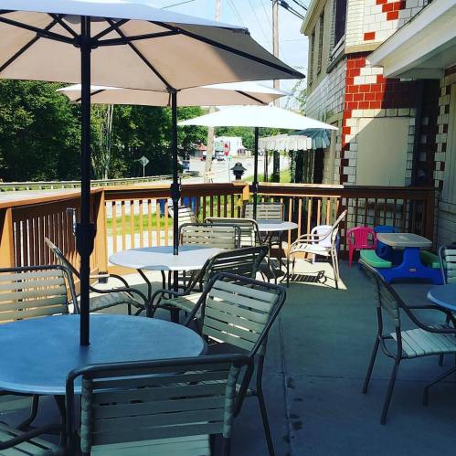 Outdoor seating area with tables and umbrellas, surrounded by greenery and a wooden railing.