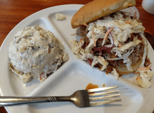 A plate with a pulled pork sandwich topped with coleslaw and a side of creamy potato salad.