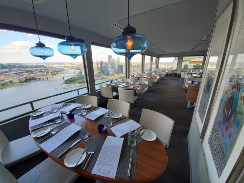 A modern restaurant with a scenic view of a river and city skyline, featuring elegant table settings and blue pendant lights.
