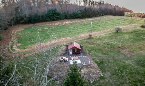 A small cabin with a red roof sits in a grassy field, surrounded by trees and a winding path.