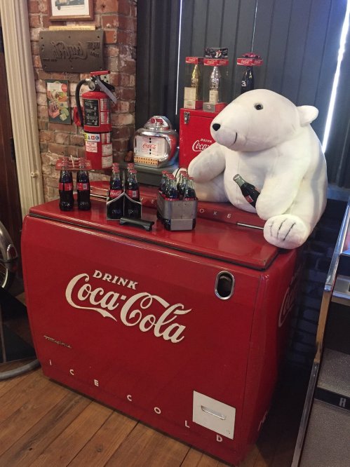 A vintage Coca-Cola cooler with bottles and a plush polar bear on top, set in a retro-themed environment.