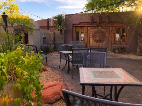 Outdoor seating area with tables, plants, and rustic buildings under a clear blue sky.