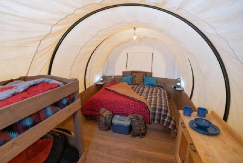 Cozy tent interior with a double bed, bunk bed, and rustic decor, featuring blue mugs and a wooden table.