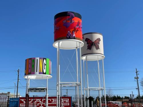 Three colorful water towers with artistic designs against a clear blue sky, featuring butterflies and bold letters.