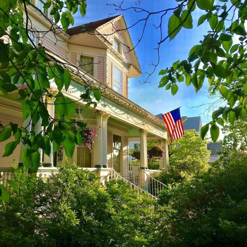 A charming house with a porch, surrounded by greenery and flowers, featuring an American flag waving in the breeze.