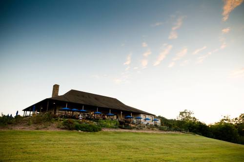 A large, rustic building with a sloped roof sits on a grassy hill under a clear sky, surrounded by trees.