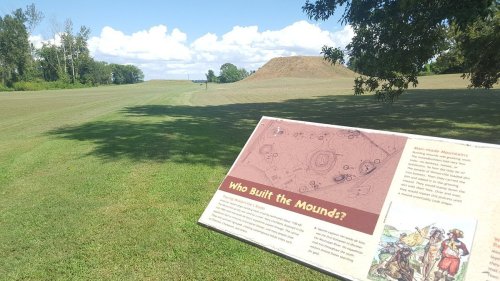 A grassy landscape with a sign about a historical mound in the background under a blue sky with clouds.