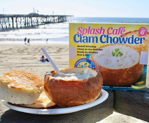 A bowl of clam chowder in a bread bowl, with a box of Splash Café clam chowder in the background, by the beach.