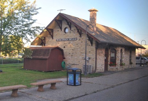 Historic brick train station with a sloped roof, surrounded by greenery and a small shed nearby.