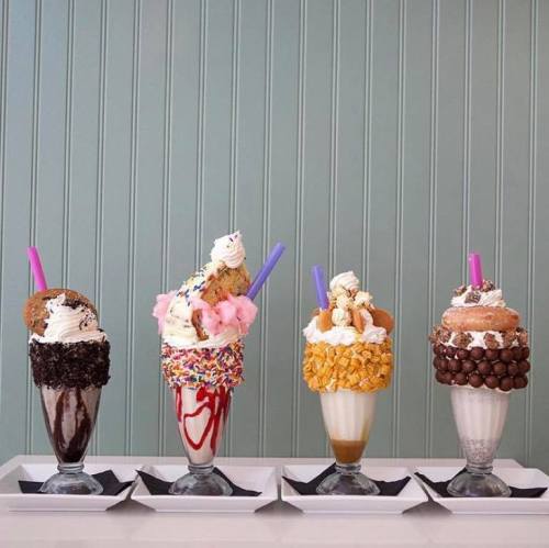 Four colorful milkshakes topped with whipped cream, cookies, candy, and cereal, served in tall glasses on a tray.
