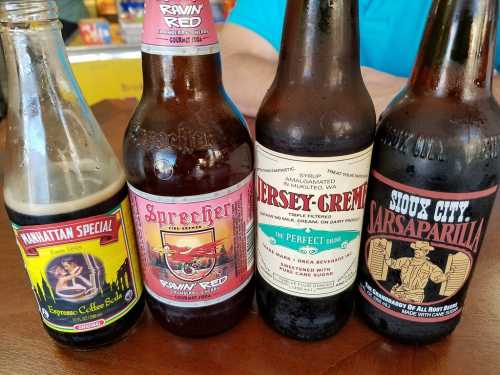 Four vintage soda bottles on a wooden table, featuring colorful labels: Manhattan Special, Sprecher, Jersey Cream, and Sioux City Sarsaparilla.