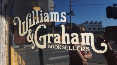 Sign on a storefront reading "Williams & Graham Booksellers," reflecting the street and nearby buildings.