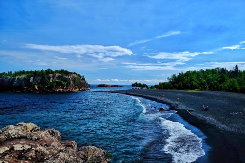 A scenic view of a beach with dark sand, blue water, and lush green trees under a bright blue sky with clouds.