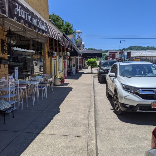 A sunny street scene featuring shops, parked cars, and trees lining the sidewalk.