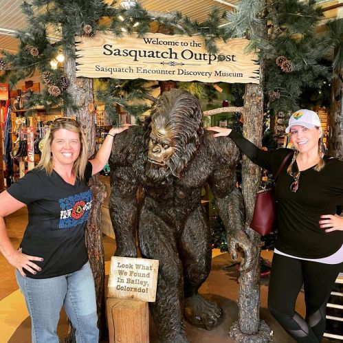 Two women pose beside a Sasquatch statue at the Sasquatch Encounter Discovery Museum, surrounded by trees and signage.