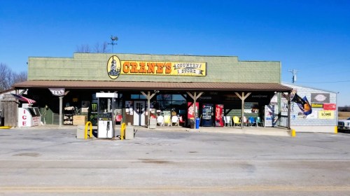 A country store with a green exterior, featuring gas pumps, outdoor seating, and various signs under a clear blue sky.