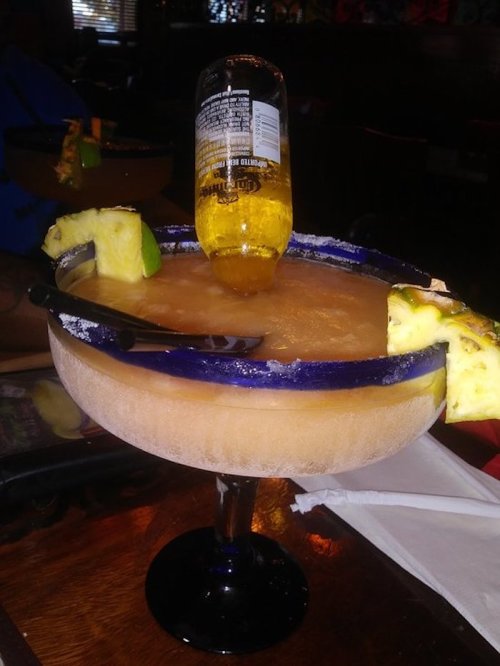 A large margarita glass with a bottle of beer inverted in it, garnished with lime and pineapple slices.