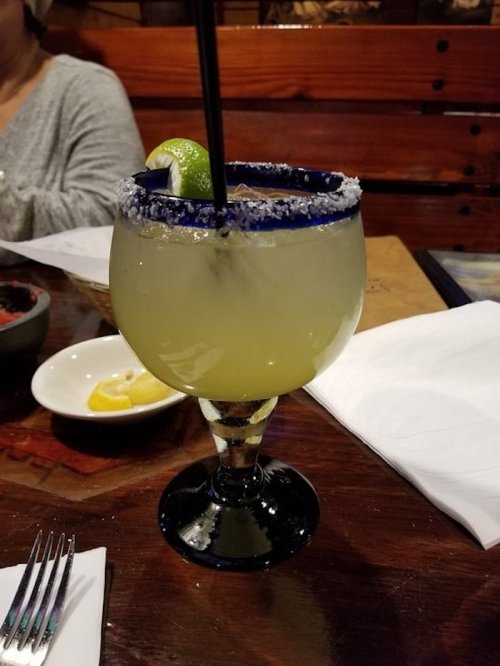 A margarita in a glass with a blue rim, garnished with a lime slice, sits on a wooden table.