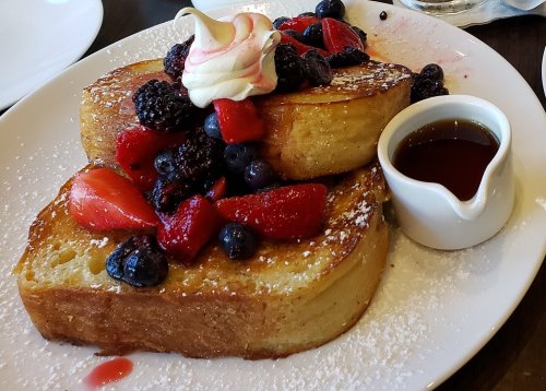 A plate of French toast topped with mixed berries and whipped cream, served with a small jug of syrup.