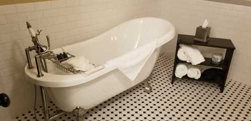 A white clawfoot bathtub with a towel draped over it, next to a black shelf holding rolled towels and a tissue box.