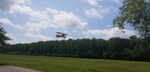 A biplane flies low over a grassy field with trees in the background under a partly cloudy sky.