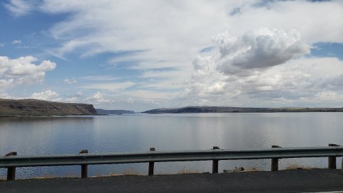 A serene lake surrounded by hills under a partly cloudy sky, with a road barrier in the foreground.
