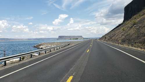 A scenic road alongside a lake, with a plateau in the distance and clouds in a blue sky.