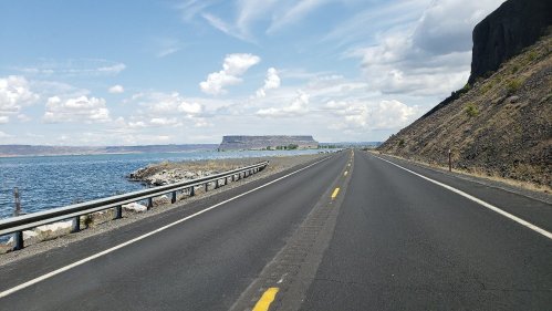 A scenic road alongside a lake, with a plateau in the distance and clouds in a blue sky.