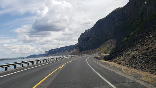 A winding road alongside a river, with steep cliffs and cloudy skies in the background.