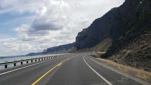 A winding road alongside a river, with steep cliffs and cloudy skies in the background.