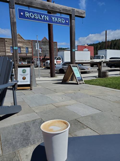 A coffee cup sits on a table in front of a sign for Roslyn Yard, with a scenic view of buildings and trees in the background.