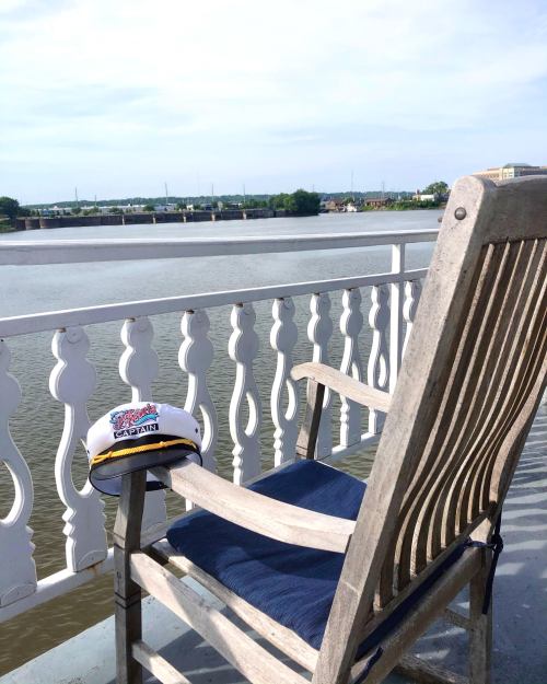 A wooden chair on a balcony overlooking a calm river, with a hat resting on the armrest.