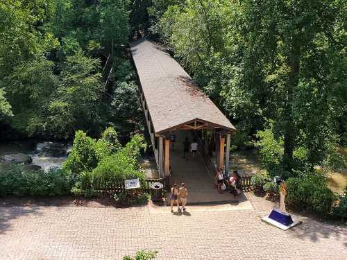 A wooden covered bridge spans a creek, surrounded by lush greenery, with people walking on the path below.