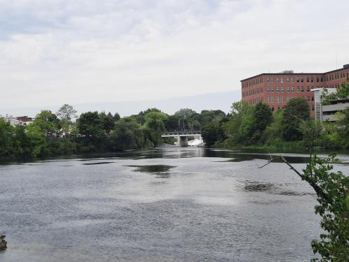 A calm river scene with trees lining the banks and a bridge in the distance, set against a cloudy sky.