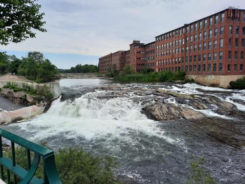A river with a waterfall flows past a large brick building, surrounded by greenery under a cloudy sky.