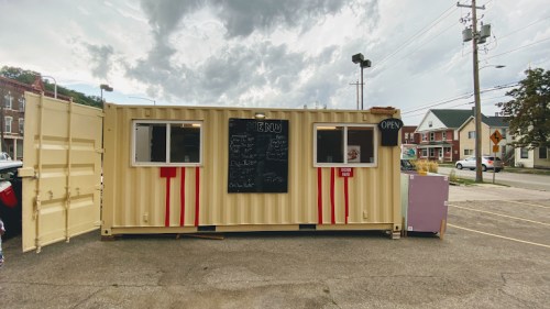 A food truck made from a shipping container, featuring a menu board and an "OPEN" sign, set against a cloudy sky.