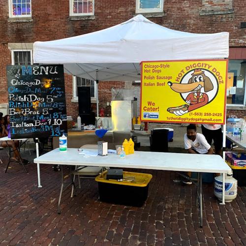 A food stand with a menu featuring hot dogs and sausages, including prices, in a brick-walled outdoor setting.