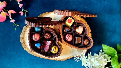 Two heart-shaped boxes of assorted chocolates on a wooden platter, surrounded by flowers and greenery.