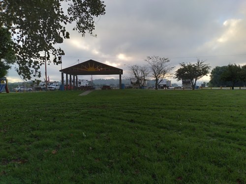 A grassy area with a pavilion in the background, under a cloudy sky, surrounded by trees and distant vehicles.