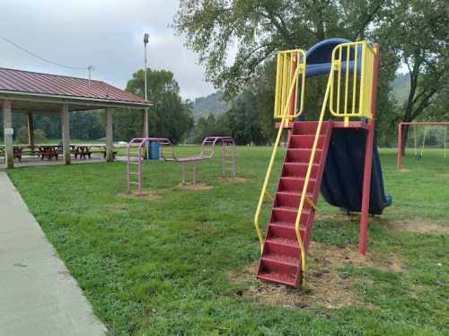 A playground with a slide, climbing bars, and swings, surrounded by grassy areas and a picnic shelter in the background.