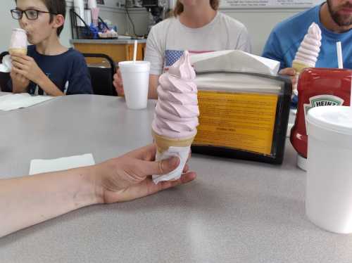 A hand holds a soft-serve ice cream cone, with people enjoying drinks and ice cream at a table in the background.