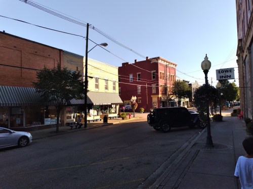 A quiet street scene with shops, trees, and a red building, bathed in warm sunlight. Cars are parked along the curb.