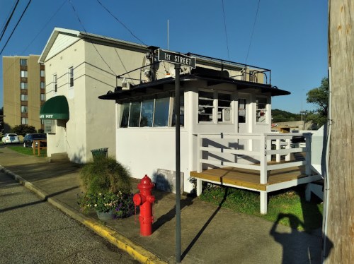 A small white building with a deck, next to a fire hydrant and a street sign for 1st Street, under a clear blue sky.