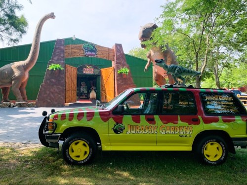 A colorful SUV parked in front of Jurassic Gardens, featuring dinosaur sculptures and a vibrant entrance.