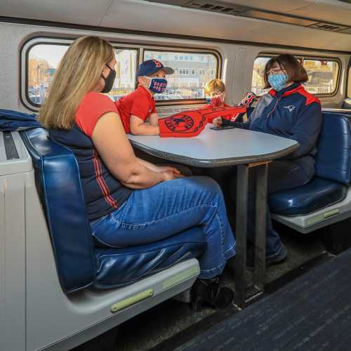 A woman and a child wearing baseball caps sit at a table in a train car, smiling and holding baseball gear.