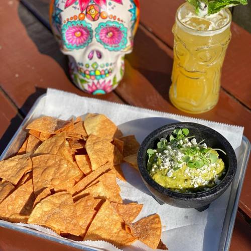 A plate of tortilla chips with guacamole, a colorful skull decoration, and a refreshing drink on a wooden table.