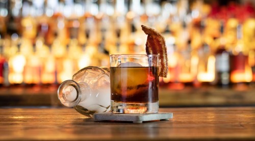 A glass of cocktail with a bacon garnish, placed on a wooden surface, with a blurred bar background.