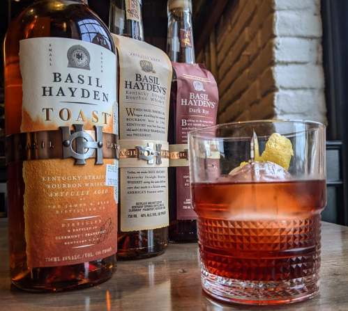 A glass of cocktail with a large ice cube, surrounded by bottles of Basil Hayden's bourbon whiskey on a wooden table.