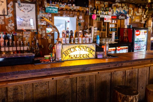 A rustic bar with a wooden counter, shelves of liquor, and a neon sign reading "Buck Snort Saloon."